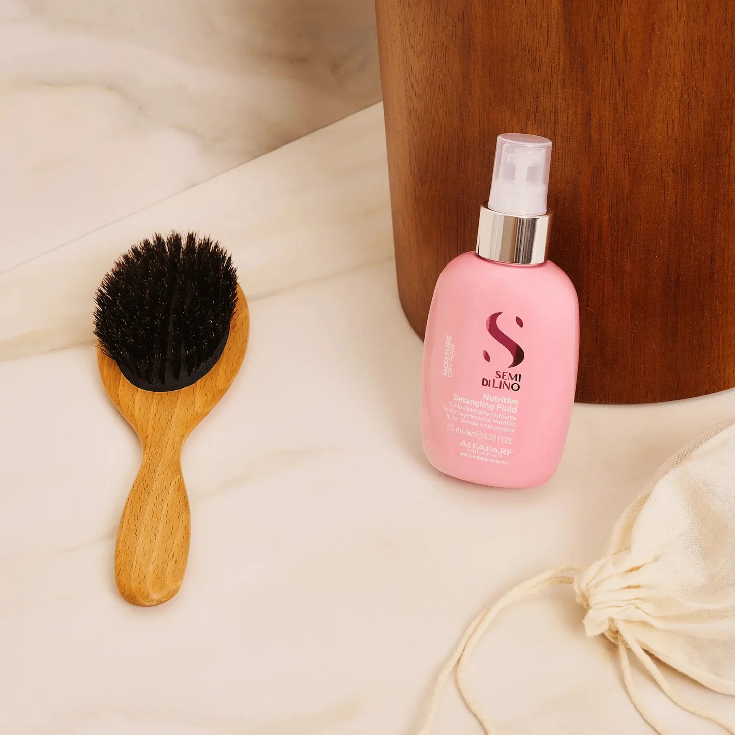 Pink Alfaparf Semi Di Lino Moisture Nutritive Detangling Fluid bottle on marble beside wooden hairbrush