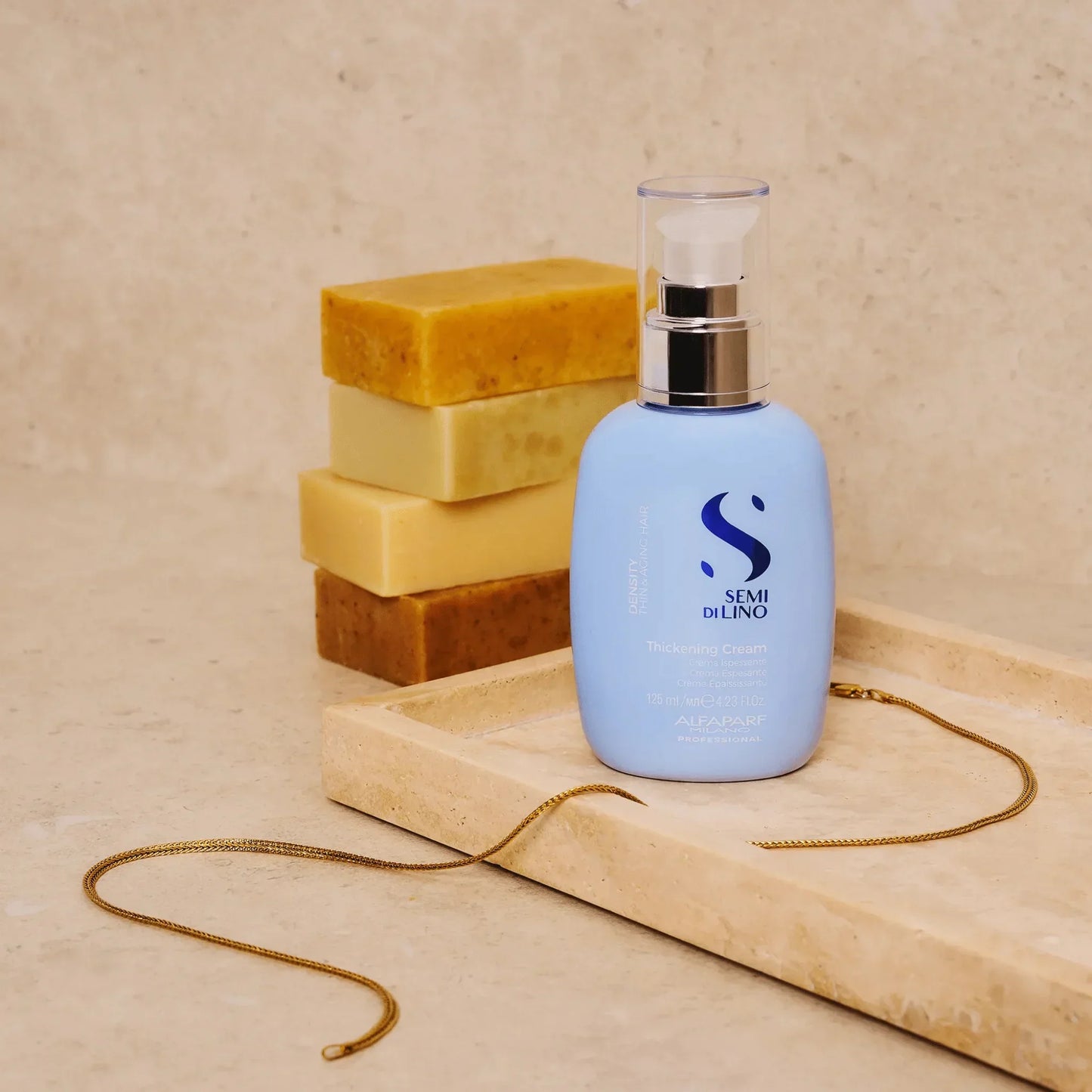 Blue bottle of thickening cream on a marble tray, stacked soap bars and gold necklace nearby.
